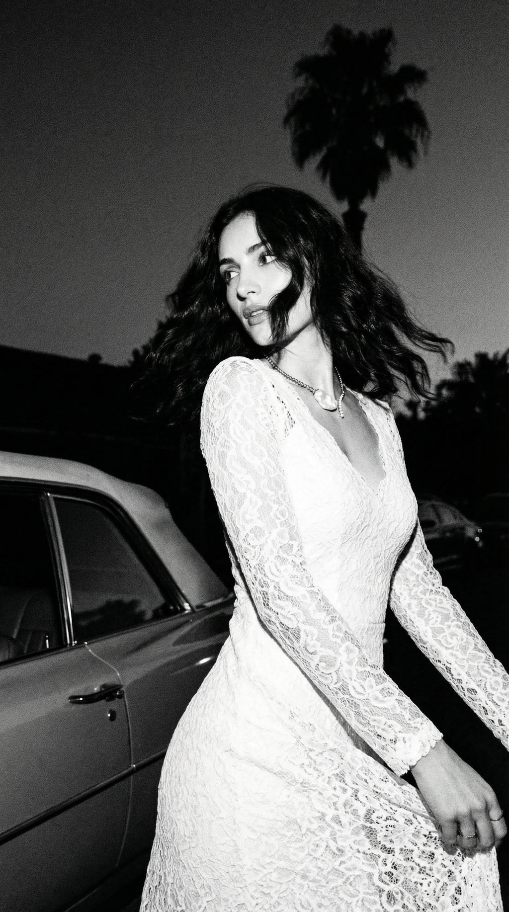 Woman in a white lace dress standing next to a car with palm trees in the background