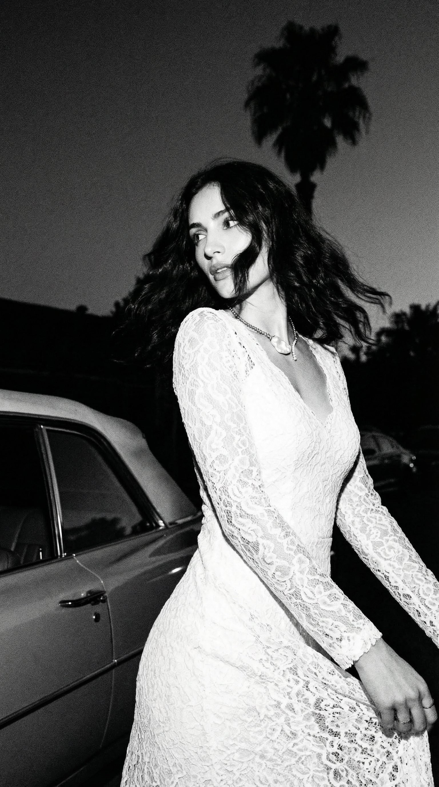 Woman in a white lace dress standing next to a car with palm trees in the background